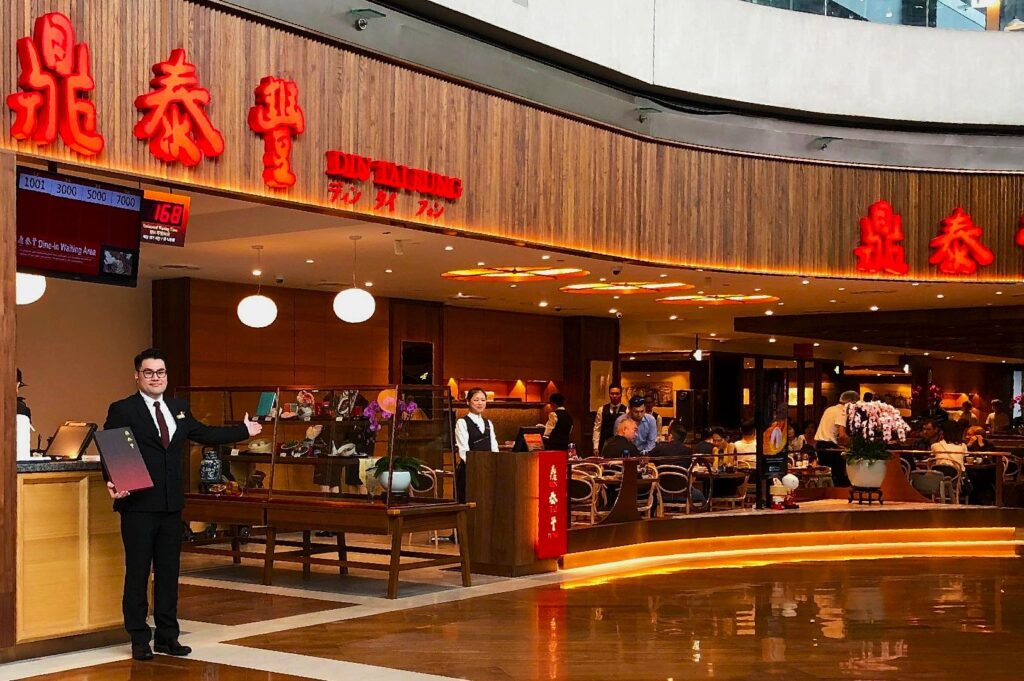 A welcoming host stands outside a vibrant, bustling restaurant with a wooden facade and bright red signage. Inside, diners chat and enjoy meals.