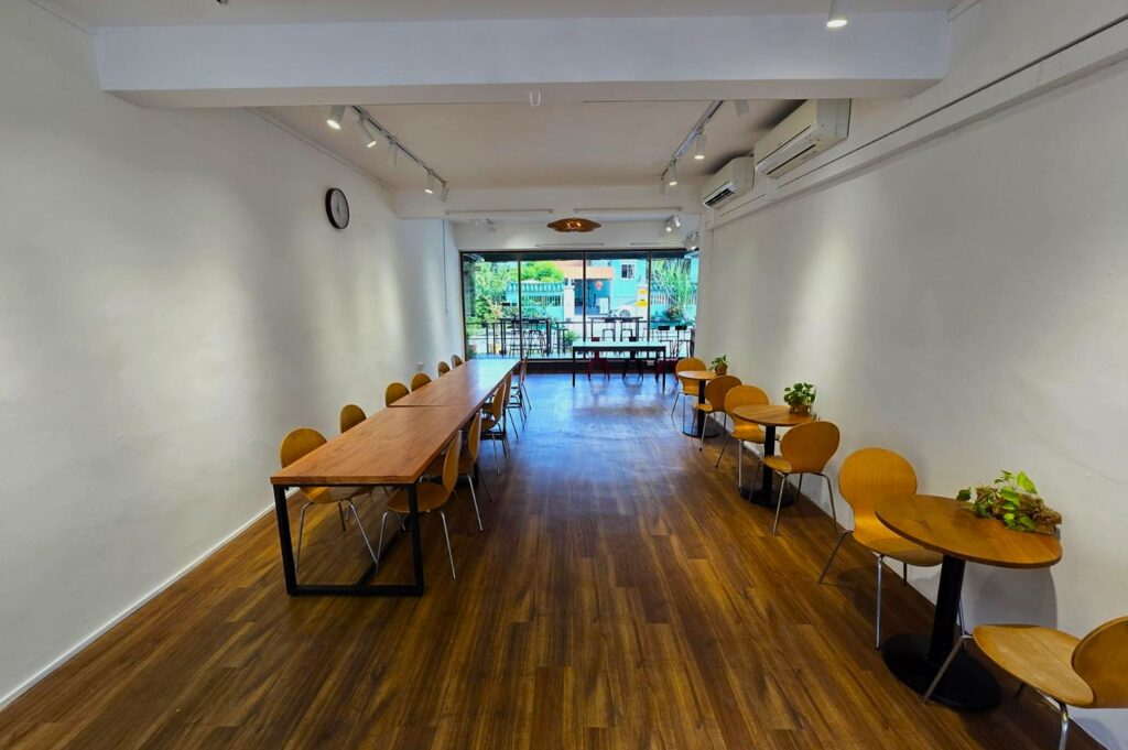 Minimalist café interior with wooden flooring and white walls. Long wooden table with chairs on left, small round tables on right, large window ahead. Cozy and bright atmosphere.