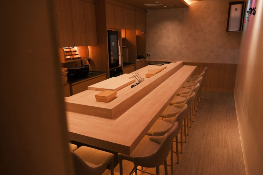 Warmly lit sushi bar interior with a long wooden counter, surrounded by chairs. Minimalist design, creating an inviting and serene atmosphere.