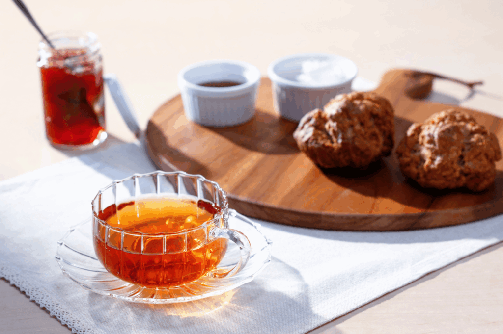 Elegant afternoon tea setup featuring a glass teacup of amber-hued tea on lace-trimmed linen, with scones and assorted condiments—jam, whipped cream, and chocolate—served on a rustic wooden board. Soft natural light evokes warmth and hospitality, capturing the refined charm of Singapore’s best tea experiences