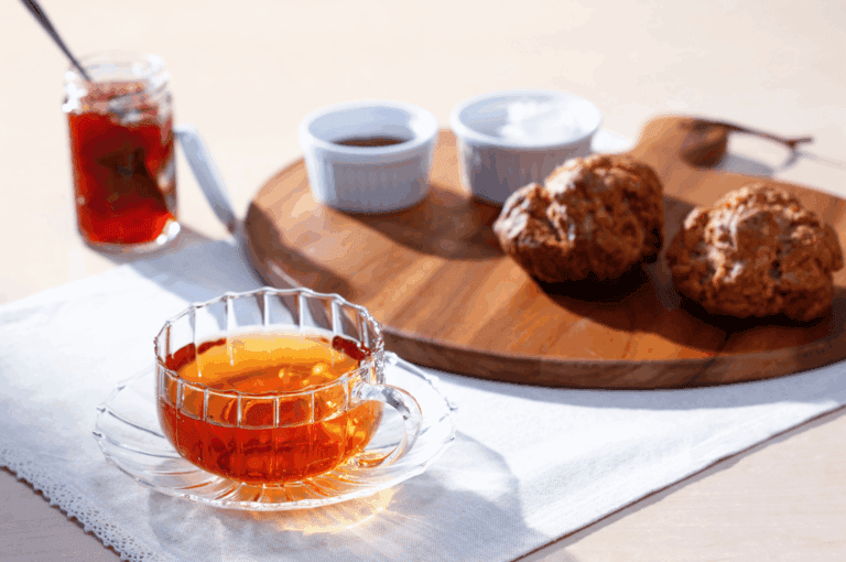 Elegant afternoon tea setup featuring a glass teacup of amber-hued tea on lace-trimmed linen, with scones and assorted condiments—jam, whipped cream, and chocolate—served on a rustic wooden board. Soft natural light evokes warmth and hospitality, capturing the refined charm of Singapore’s best tea experiences