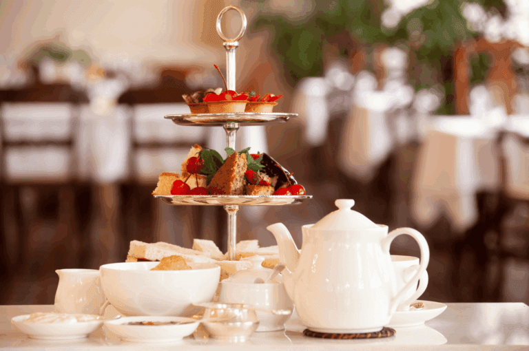Refined afternoon tea presentation in an upscale Singapore dining room, featuring a three-tiered stand with cherry-topped pastries, mint-garnished cakes, and crustless finger sandwiches. Surrounded by white porcelain teaware and condiments, the elegant setup evokes timeless hospitality and cultural sophistication.