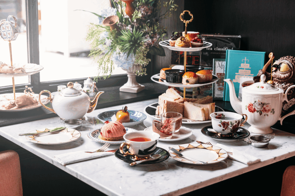 Luxurious afternoon tea spread on a marble table by the window, featuring floral porcelain teaware with gold accents, tiered stands of pastries, scones, and cakes, plus sandwiches and pink mousse. A vase of flowers and stacked books complete the cozy, vintage ambiance—evoking Singapore’s elegant tea rituals and refined hospitality.