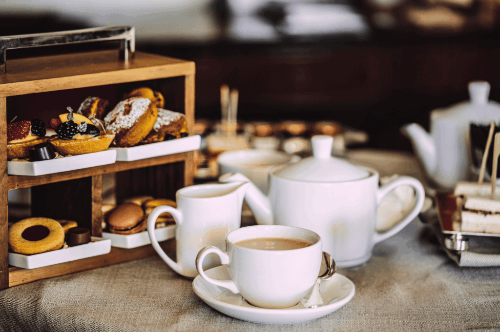 Cozy afternoon tea scene with a white porcelain teapot, milk jug, and teacup beside a wooden tiered stand of fruit-topped tarts, powdered cakes, cookies, and macarons. Set on a beige tablecloth with soft lighting, the elegant spread reflects Singapore’s warm hospitality and refined tea traditions.