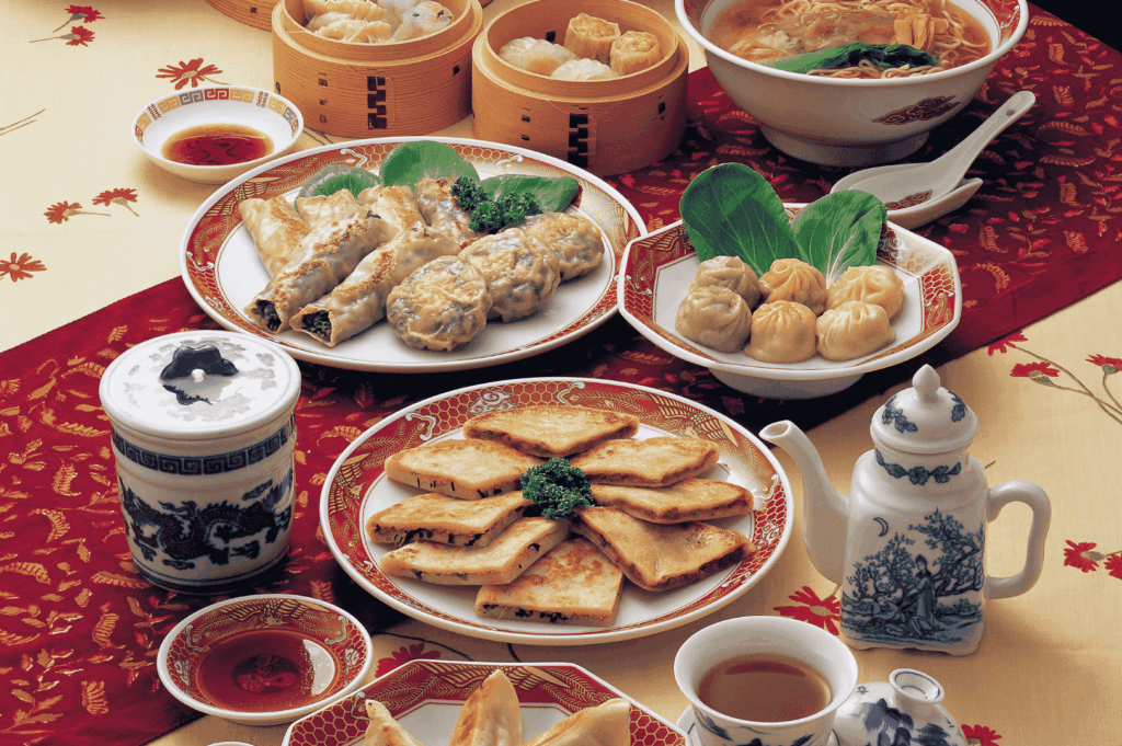 A vibrant overhead view of a traditional Chinese meal spread on a red-and-gold patterned tablecloth, showcasing regional diversity beyond Cantonese cuisine. The table features bamboo steamers filled with assorted dumplings—steamed and pan-fried—alongside spring rolls, noodle soup with leafy greens, and plates of bok choy-accompanied dumplings. Ornate red-and-white porcelain dishes and a classic Chinese teapot evoke cultural richness, while the variety of textures and preparations hints at Sichuan, Shanghainese, and Northern Chinese influences within Singapore’s evolving culinary landscape.