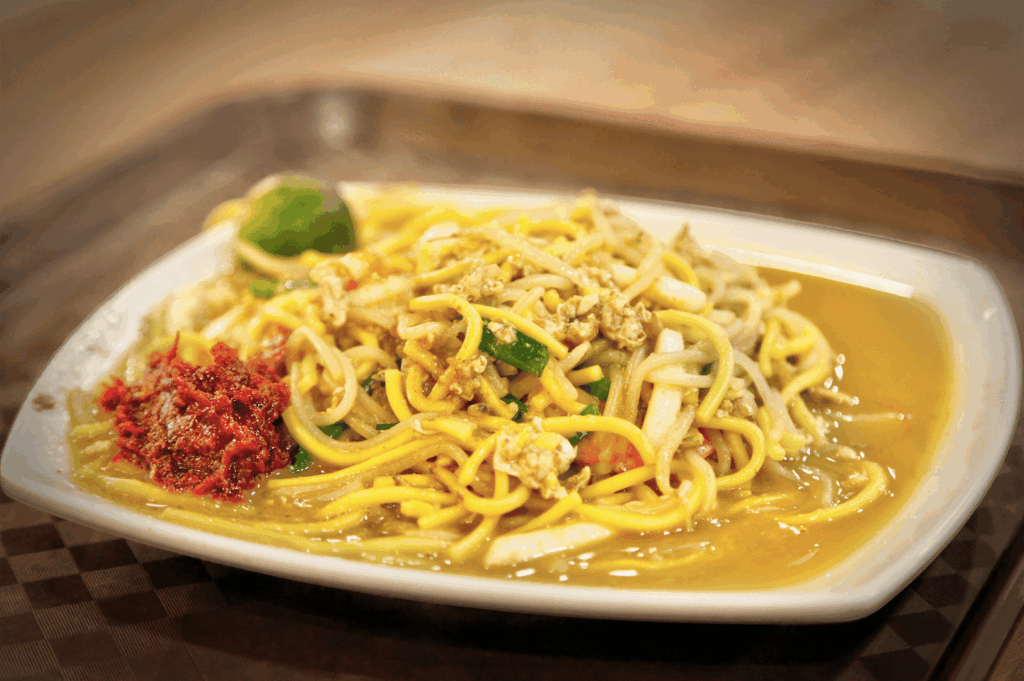 A plate of yellow noodles drenched in thick, savory gravy, garnished with bean sprouts, sliced green chili, and hints of seafood or meat—likely a Singapore-style Hokkien Mee. A dollop of red chili paste and a halved lime rest on the side, inviting customization. Presented on a white rectangular plate atop a brown checkered tray, the dish evokes Southeast Asian comfort food rooted in Fujianese culinary heritage, expanding the narrative of Chinese regional flavors in Singapore beyond Cantonese norms.