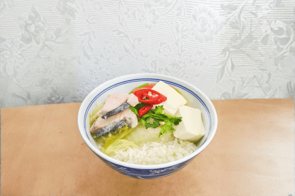 A ceramic blue-and-white bowl filled with white rice and topped with cooked salmon, tofu cubes, napa cabbage, red chili slices, and fresh cilantro—an earthy, nourishing composition that blends protein, spice, and greens. Set against a floral-patterned wall and wooden surface, the dish evokes homestyle warmth and quiet refinement. Its balanced flavors and visual restraint suggest Teochew or Hakka influences, expanding the story of Chinese regional cuisines thriving in Singapore beyond the dominant Cantonese lens.