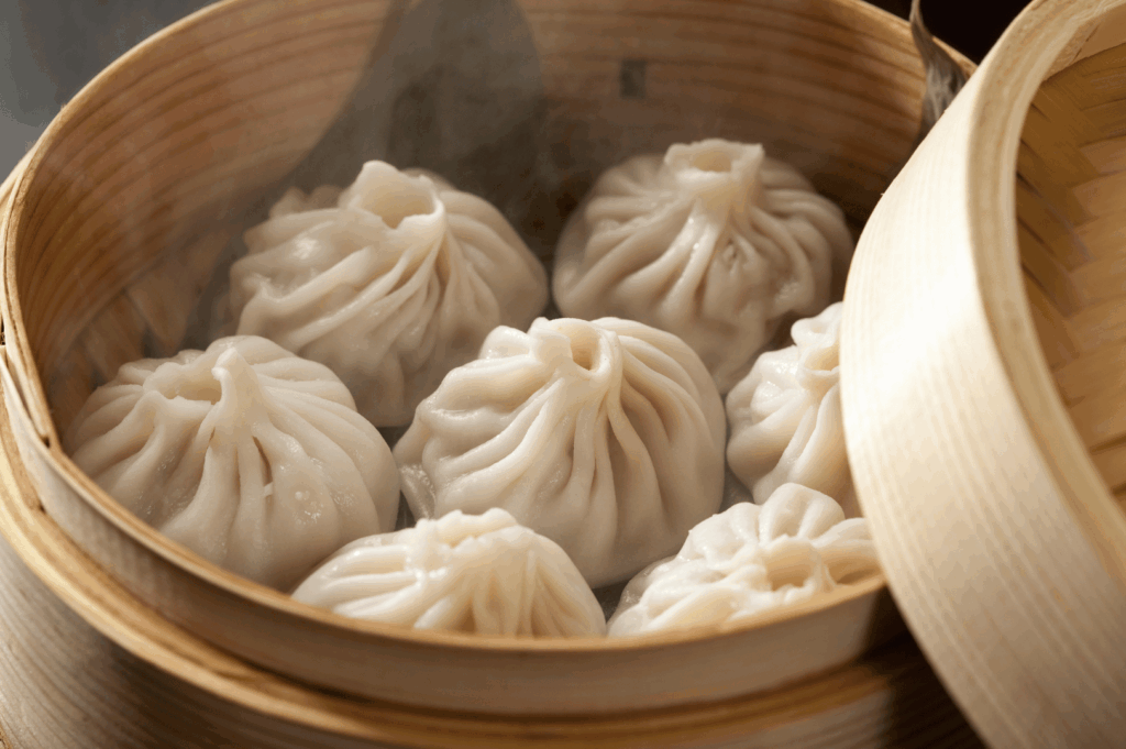 A bamboo steamer filled with freshly steamed xiao long bao, their pleated tops glistening as steam rises from the open lid. The soup dumplings are arranged with care, each one showcasing the delicate craftsmanship of Northern Chinese culinary tradition. The warm, tactile texture of the bamboo contrasts with the soft dumplings, evoking intimacy and ritual. This image anchors the editorial’s exploration of Chinese regional cuisines in Singapore, spotlighting the quiet elegance of non-Cantonese traditions like Shanghainese soup dumplings.