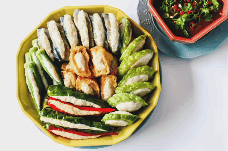 A vibrant platter of assorted stuffed vegetables and dumplings, artfully arranged on a yellow dish with a blue base. Bitter melon, green chili, and other vegetables are filled with white fish paste or tofu, accompanied by golden-brown fried tofu and translucent-skinned dumplings. A red bowl of dipping sauce garnished with cilantro and sliced red chili adds brightness and heat. This image celebrates the culinary precision and bold textures of Hakka and Teochew traditions, enriching Singapore’s Chinese food narrative beyond Cantonese staples.