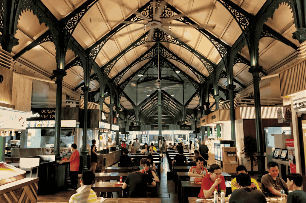 Busy Singapore hawker center with ornate iron-beam ceiling, food stalls lining both sides, and groups gathered at wooden tables — showcasing the city’s vibrant communal dining culture.