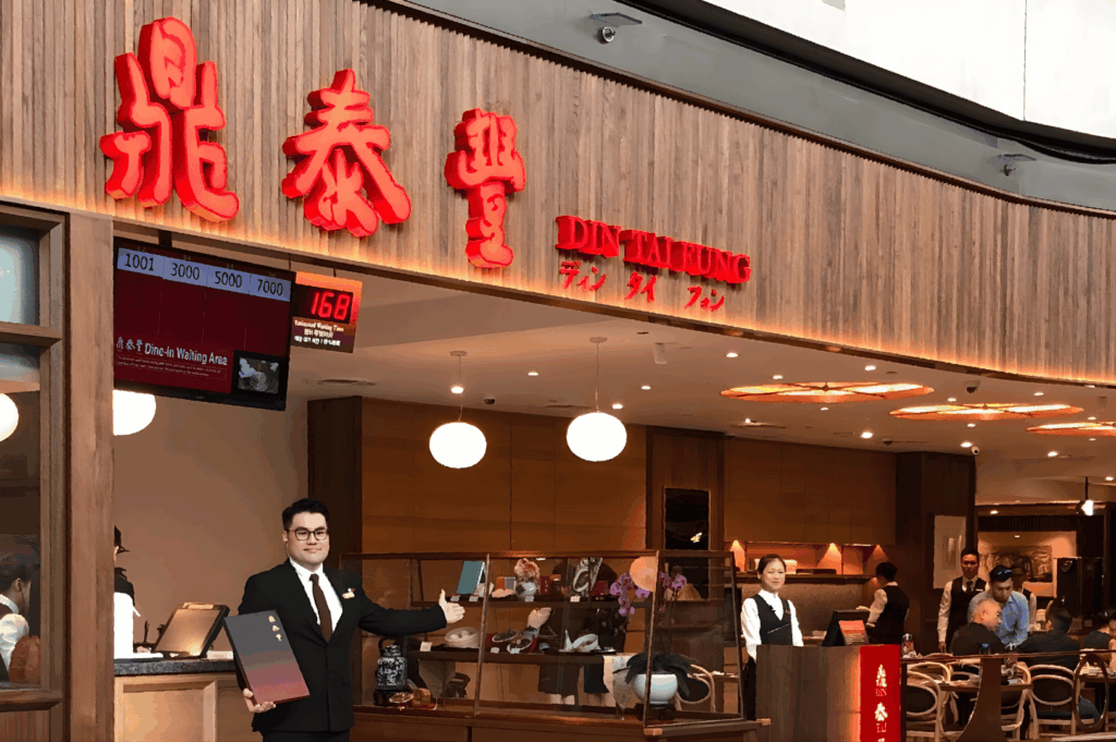 Exterior of Din Tai Fung in Singapore with red signage, digital queue display, and a suited staff member holding menus — reflecting the popularity and polished service of this iconic group dining destination.