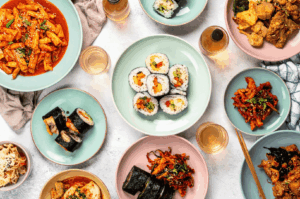A vibrant overhead shot of a contemporary Korean meal spread in Singapore, showcasing sliced kimbap rolls, spicy tteokbokki garnished with sesame seeds and scallions, crispy fried chicken, stir-fried pork or squid, and assorted banchan including kimchi and pickled vegetables. The dishes are plated on pastel-toned ceramics, surrounded by glasses of light-colored tea or soju, reflecting the playful yet refined aesthetic of modern Korean dining. This image captures the evolving Korean culinary landscape in Singapore—where tradition meets innovation in color, texture, and communal spirit.