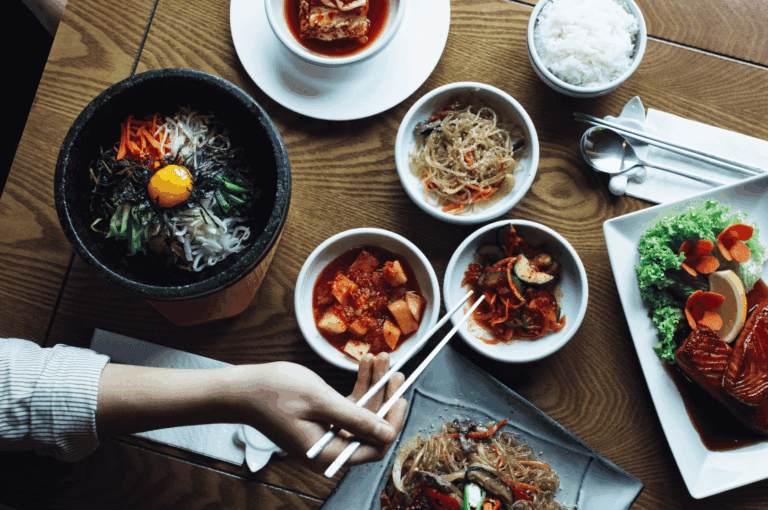 A warm, inviting tablescape of a traditional Korean meal in Singapore, featuring a hot stone bowl of bibimbap with vibrant vegetables, seaweed, and a raw egg yolk; a bubbling bowl of kimchi stew; japchae noodles being lifted by chopsticks; sliced grilled meat with lettuce garnish; and an array of banchan including kimchi, kkakdugi, and seasoned vegetables. The composition evokes the communal spirit and sensory richness of Korean dining, reflecting how heritage dishes remain central to Singapore’s evolving Korean food scene.