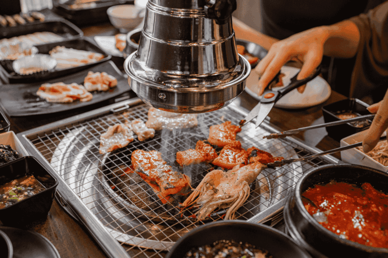 Dynamic scene of Korean barbecue dining in Singapore, with marinated meats and seafood sizzling over a tabletop grill beneath a stainless steel exhaust hood. Two diners actively cook—one using scissors to cut meat, the other handling tongs—surrounded by trays of raw ingredients, vegetables, and dipping sauces. The image captures the communal energy and hands-on experience of Korean barbecue, reflecting its immersive appeal and growing influence in Singapore’s evolving Korean food culture.