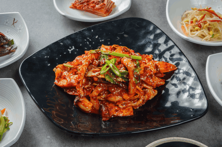 A visually striking plate of Ojingeo Bokkeum—spicy stir-fried squid coated in a vibrant gochujang-based sauce—garnished with sesame seeds and sliced green chili, served on a black square plate. Surrounding banchan dishes include kimchi, seasoned bean sprouts, and assorted marinated vegetables, all arranged on white plates. This image captures the bold flavors and fiery elegance of Korean cuisine, reflecting its continued evolution and cultural resonance in Singapore’s dynamic food scene.