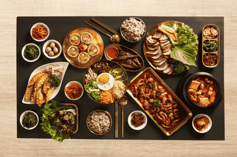 Expansive Korean meal spread featuring bibimbap topped with a fried egg, sliced pork belly with lettuce wraps and sauces, spicy stir-fried squid, kimchi stew with tofu, golden tray of kimbap, dumplings, grilled fish, and a variety of banchan including seasoned greens, pickled vegetables, and quail eggs. Two bowls of multigrain rice anchor the setup. This image celebrates the abundance and diversity of Korean cuisine, reflecting its deep cultural roots and evolving presence in Singapore’s vibrant food scene.