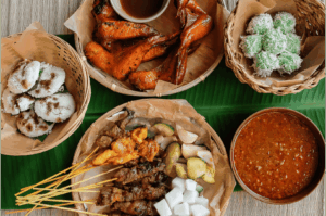 A vibrant spread of traditional Malay dishes arranged on a banana leaf, featuring grilled chicken wings, coconut-coated glutinous rice balls, palm sugar-topped rice cakes, assorted satay skewers, and a rich peanut dipping sauce—capturing the rustic essence of communal dining and the foundational flavors that continue to inspire modern interpretations of Malay cuisine.