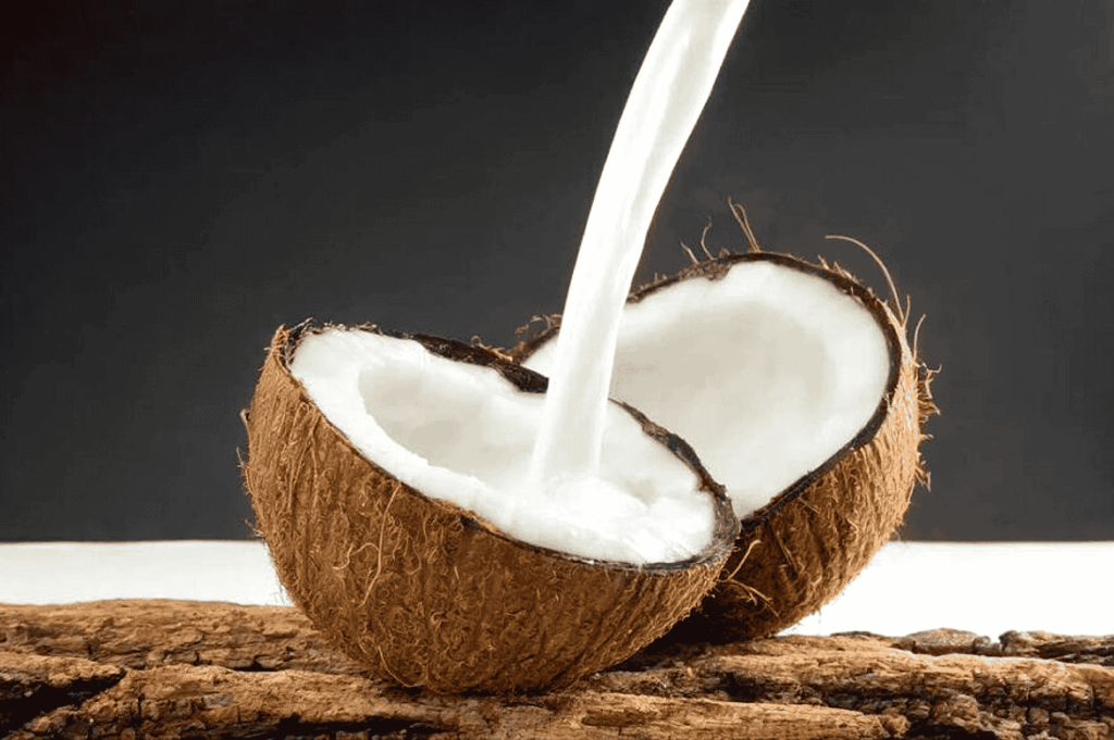 A halved coconut on a rustic wooden surface, with fresh coconut milk cascading into its shell against a dark, moody backdrop—symbolizing the elemental purity and enduring role of coconut in Malay cuisine, from ancestral recipes to modern reinterpretations.