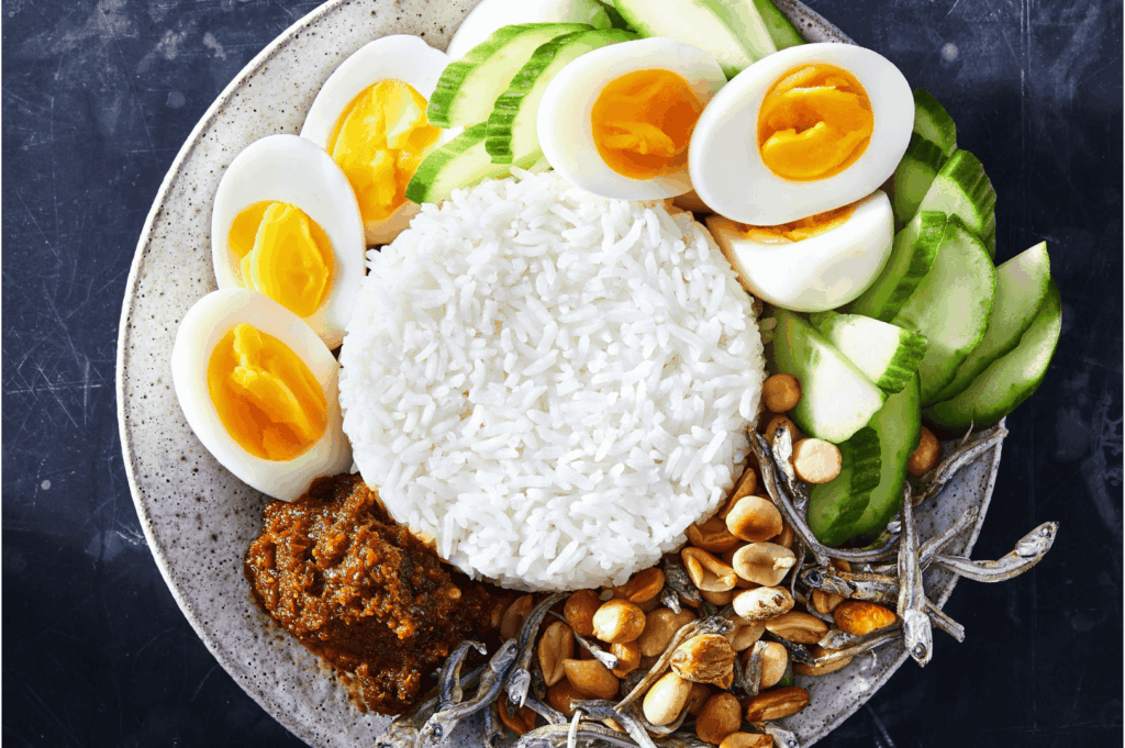 A visually balanced plate of Nasi Lemak featuring fragrant coconut rice surrounded by sambal, crispy anchovies, roasted peanuts, hard-boiled eggs, and fresh cucumber slices—embodying the harmony of flavors, textures, and cultural symbolism that anchor traditional Malay cuisine and continue to influence its modern evolution.