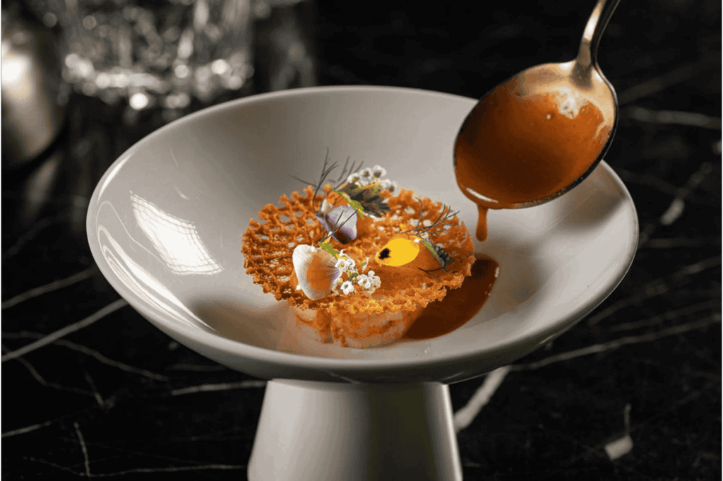 A high-concept reinterpretation of Malay flavors, plated in a white pedestal bowl with a golden crisp base, edible flowers, and delicate garnishes—elevated by a dramatic pour of reddish-brown sauce, evoking the artistry and innovation driving modern Malay cuisine into the realm of fine dining.