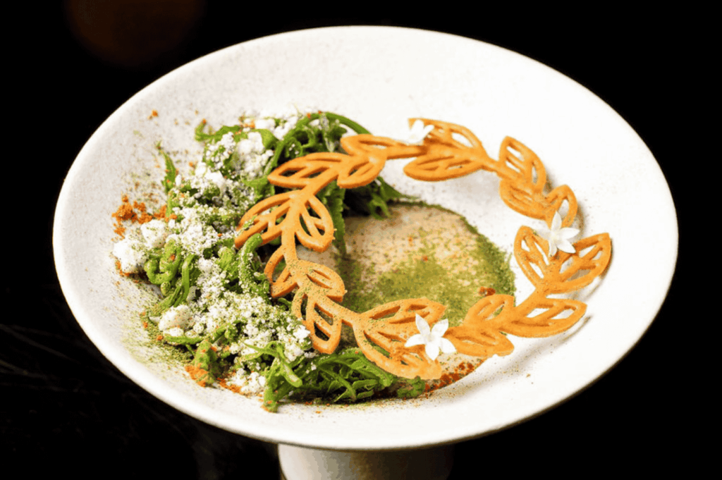 An artfully plated modern Malay dish on a white pedestal plate, featuring a wreath of golden crisps adorned with white edible flowers, vibrant green vegetables dusted with spice, and a creamy herb-infused center—showcasing the meticulous craftsmanship and visual poetry of contemporary Malay gastronomy.
