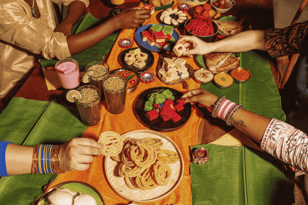 A vibrant overhead view of a festive Indian snack spread in Singapore, 2025, showcasing jalebi, barfi, gulab jamun, and colorful drinks arranged on banana leaves and bright cloth. Multiple hands reach in from the edges, capturing the communal joy of celebratory dining. Small oil lamps (diyas) dot the table, evoking Diwali warmth and the sensory richness of regional Indian food traditions. The image reflects the diversity and emotional resonance of South Asian culinary rituals in Singapore’s evolving foodscape.