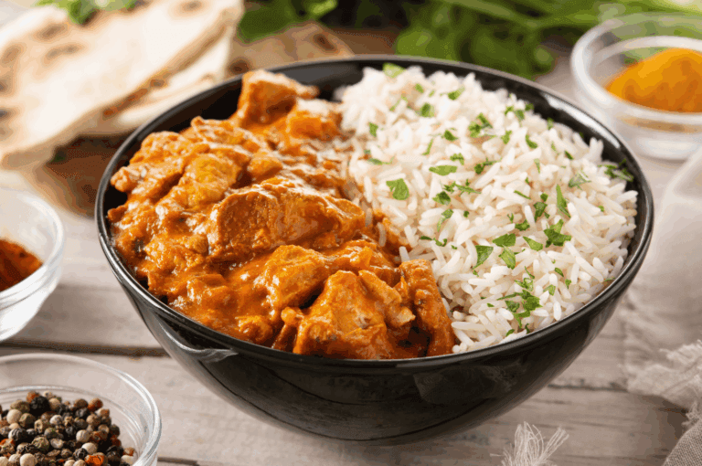 A cinematic close-up of chicken tikka masala and white rice in a black bowl, garnished with fresh herbs and surrounded by vibrant Indian spices—turmeric, chili powder, and peppercorns—on a rustic surface. A piece of flatbread and sprigs of green herbs frame the composition, evoking the sensory depth of North Indian cuisine. The warm orange sauce and rich textures highlight the dish’s comforting appeal, anchoring the narrative of regional Indian flavors in Singapore’s 2025 dining scene.