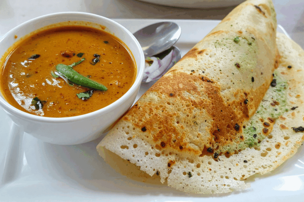 A crisp South Indian dosa served with a bowl of sambar on a white rectangular plate, garnished with curry leaves and green chili. The dosa reveals a hint of green chutney inside, while a spoon and slice of onion add tactile detail. The clean composition and warm tones evoke the comforting simplicity of traditional South Indian breakfasts, anchoring the diversity of Indian regional cuisine in Singapore’s 2025 culinary guide.