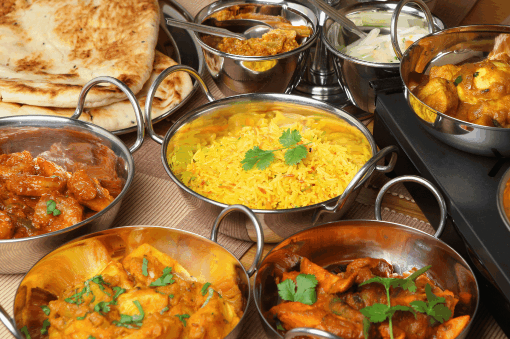An abundant spread of Indian curries—chicken, paneer, and mixed vegetables—served in traditional metal bowls and garnished with fresh cilantro, alongside turmeric-infused yellow rice, naan bread, and a crisp cucumber-onion salad. The rich colors and layered textures evoke the sensory complexity of regional Indian cuisine, celebrating spice, warmth, and hospitality. This image anchors the editorial’s exploration of culinary diversity in Singapore’s 2025 Indian food scene, highlighting communal abundance and cultural depth.