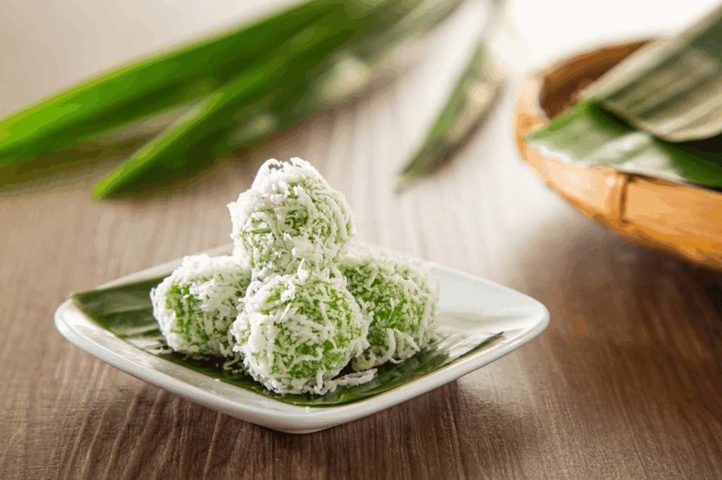 Close-up of ondeh-ondeh—green pandan glutinous rice balls coated in shredded coconut—stacked on a small white plate with banana leaf, highlighting a classic Singapore sweet.