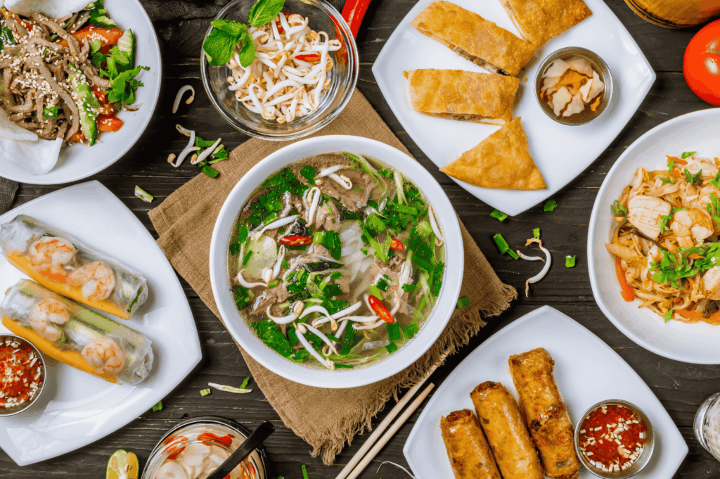 A vibrant spread of Vietnamese street food in Singapore, featuring a steaming bowl of beef pho with fresh herbs and chili at the center, surrounded by shrimp spring rolls, golden fried egg rolls, grilled chicken with herbs, stir‑fried noodles, and pickled vegetables on a dark wooden table.