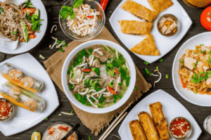 A vibrant spread of Vietnamese street food in Singapore, featuring a steaming bowl of beef pho with fresh herbs and chili at the center, surrounded by shrimp spring rolls, golden fried egg rolls, grilled chicken with herbs, stir‑fried noodles, and pickled vegetables on a dark wooden table.