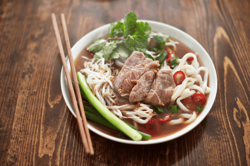 Close-up of a steaming bowl of Vietnamese beef pho in Singapore, with rice noodles, tender beef slices, fresh cilantro, bean sprouts, red chili, and green onions in clear broth, served with wooden chopsticks on a rustic wooden table.