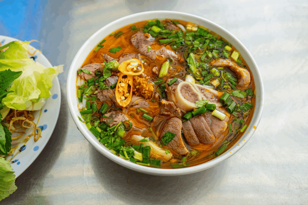 Spicy Vietnamese Bún bò Huế noodle soup in Singapore, with tender beef slices, marrow bone, chili oil broth, and fresh herbs, served alongside a plate of lettuce, mint, and bean sprouts for an authentic street food experience.