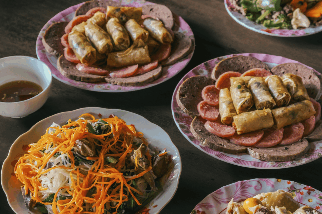 Assorted Vietnamese street food in Singapore, featuring fried spring rolls layered over pork sausage and luncheon meat on rice paper, served with fish sauce dip, alongside a vermicelli noodle salad with herbs, dumplings, and fresh vegetable sides.