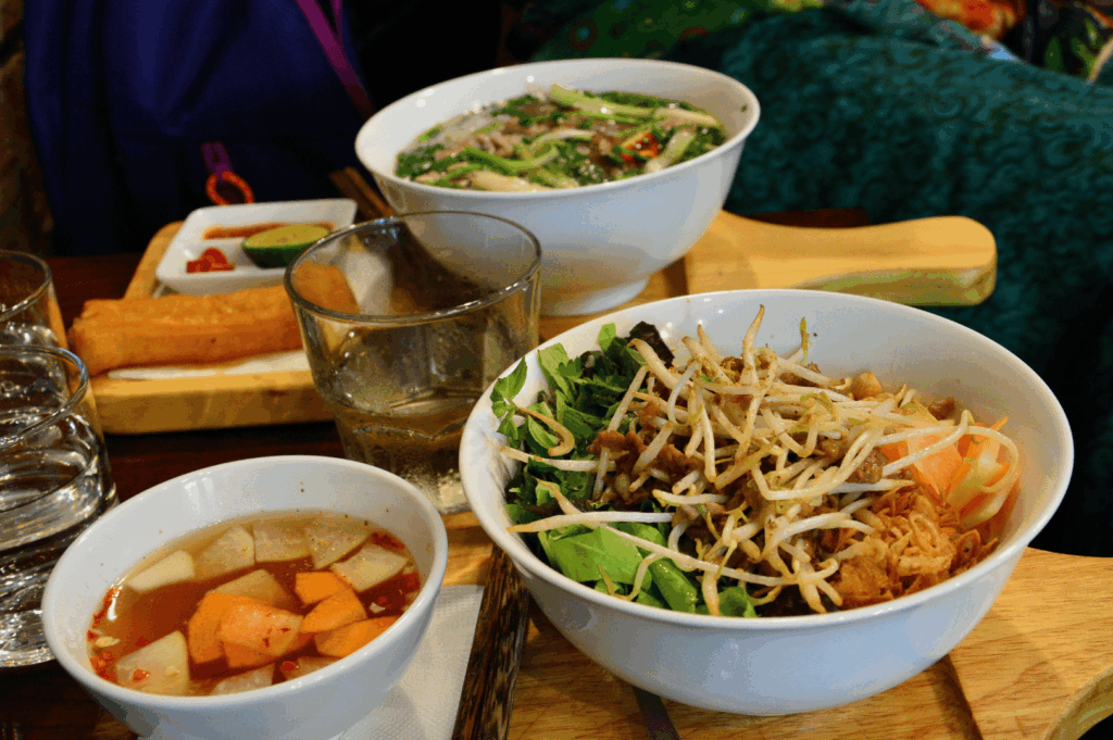 Vietnamese street food meal in Singapore with a vermicelli noodle bowl topped with grilled meat, bean sprouts, and herbs, served alongside dipping sauce with pickled vegetables, a steaming bowl of beef pho, a fried spring roll, and condiments on a restaurant table.