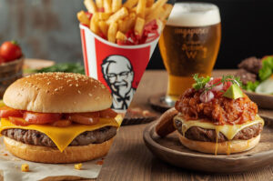 Split image showing a cheeseburger with tomato slices and a KFC bucket of fries on the left, and a messy burger with cheese, relish, and a glass of beer on the right.