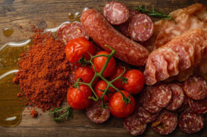 Overhead shot of a charcuterie arrangement on a wooden board, featuring sliced salami and cured meats, grape tomatoes on the vine, a pile of red paprika powder, and drizzles of olive oil.