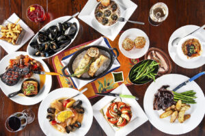 Overhead view of a wooden table completely covered with a classic French bistro dinner, including mussels, escargots, steak with frites, fish, French onion soup, and wine glasses.