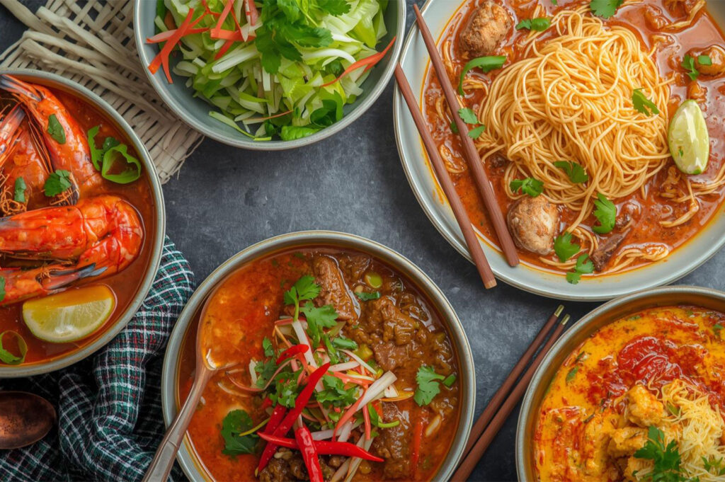 An overhead arrangement of multiple vibrant Thai dishes, including a rich prawn curry (likely Tom Yum/Kha), a bowl of beef curry, and a dish of noodles with meatballs in a red sauce, alongside a fresh side salad.