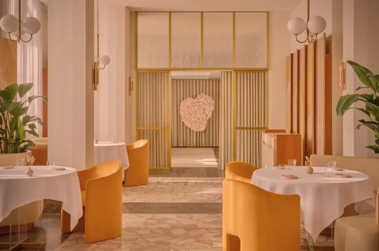 Luxurious restaurant interior with warm tones featuring round tables with white tablecloths, mustard chairs, plants, and a heart art piece on the wall.