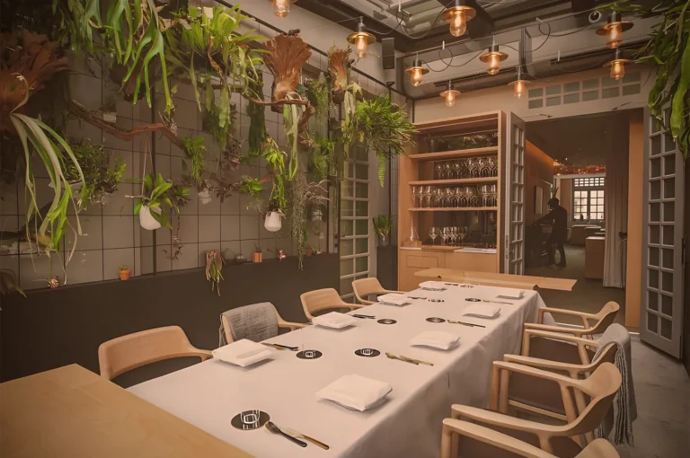 Stylish dining room with a long table set for eight. Walls adorned with lush hanging plants and warm pendant lights, creating a cozy, inviting atmosphere.