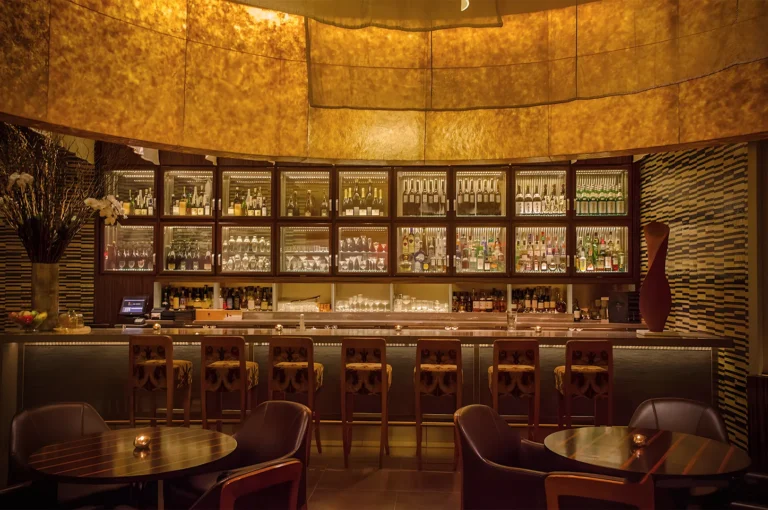 Elegant bar interior with warm lighting, wooden stools at the counter, and shelves stocked with bottles and glasses. Cozy, inviting atmosphere.