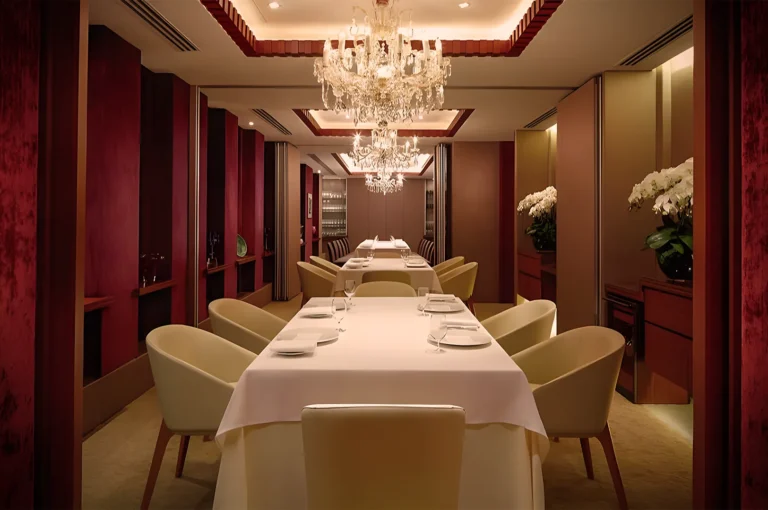 Elegant restaurant interior with chandeliers, white tablecloths, and plush chairs. Burgundy walls and potted orchids create a luxurious atmosphere.