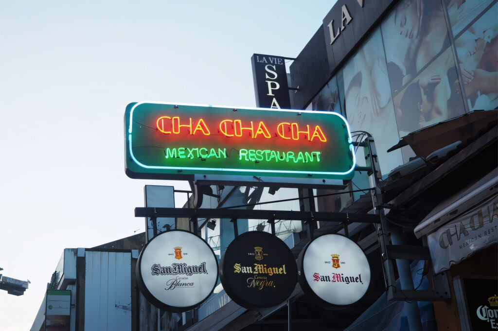 Exterior view of a Mexican restaurant with a bright red and green neon sign reading "CHA CHA CHA MEXICAN RESTAURANT," featuring three circular signs for San Miguel beer brands below it, against a clear sky.