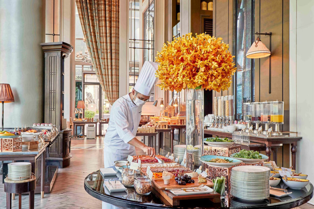 A chef in a white uniform and mask arranging food on a lavish breakfast or brunch buffet table in a grand, high-ceilinged luxury restaurant with large windows and a vase of orange flowers.