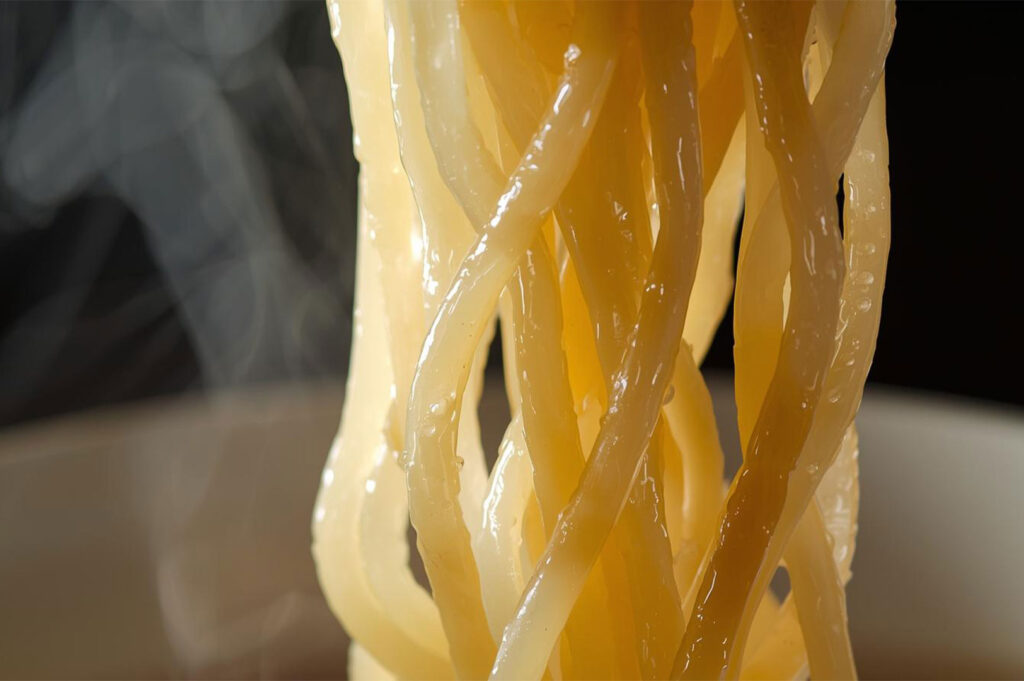 Extreme close-up macro shot of thick, glistening Japanese noodles (likely udon or thick ramen noodles) being lifted, showing their texture and wisps of steam against a dark background.
