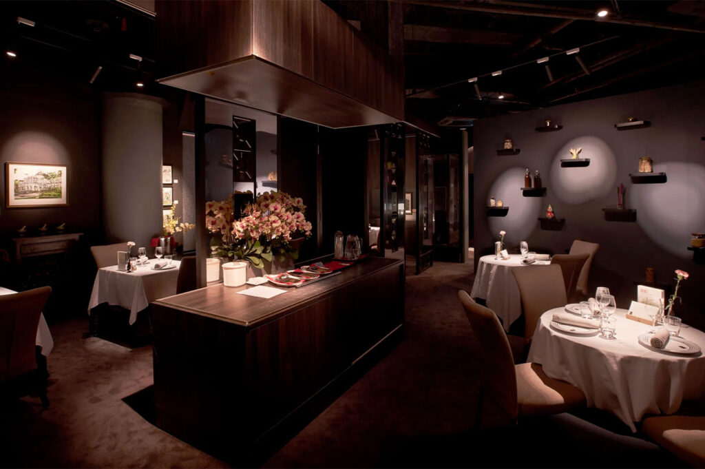 Interior view of a luxurious, dimly lit private dining room or high-end restaurant with a dark color palette. Features small, white cloth-covered tables, plush seating, a central floral arrangement, and spotlights highlighting decorative elements on the walls.
