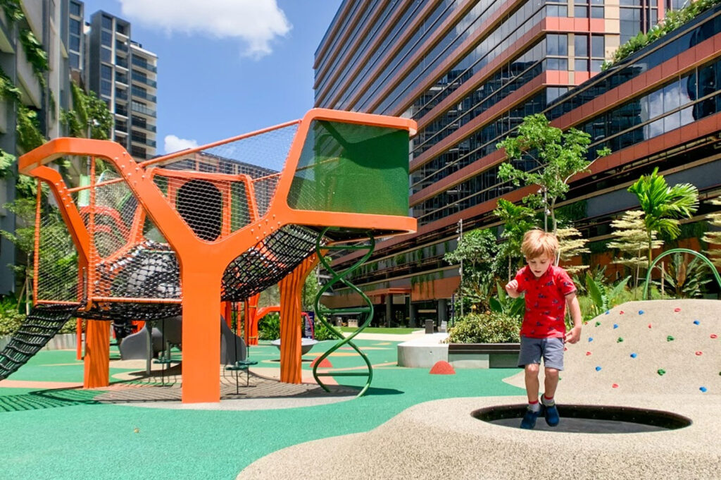 A young boy running on an outdoor playground with a modern orange climbing structure and soft landing turf, surrounded by modern residential or commercial buildings.