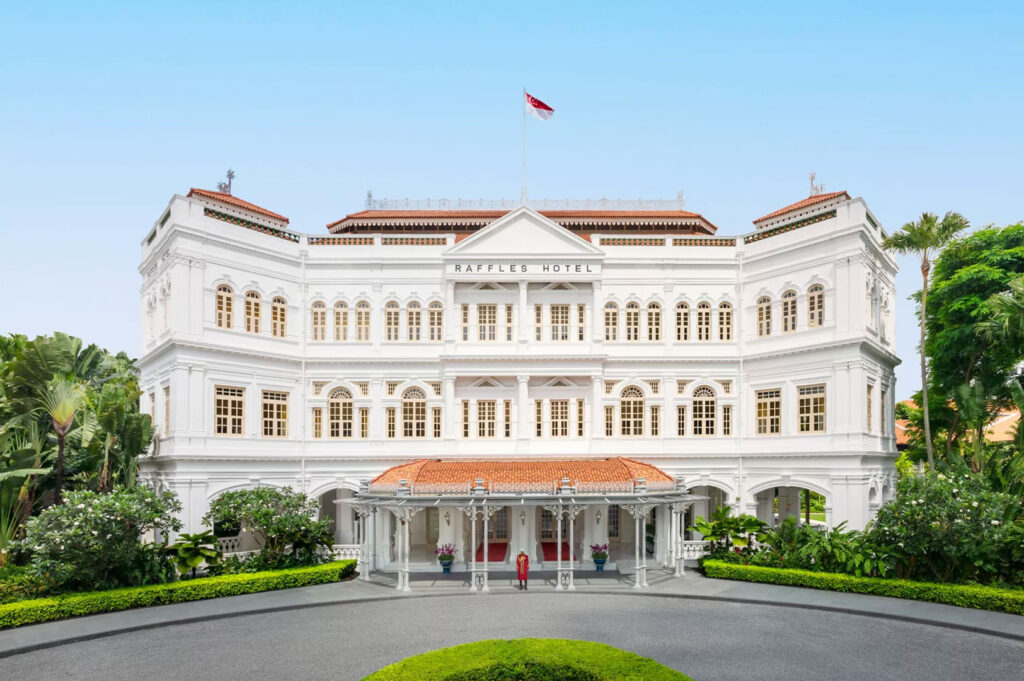 The grand, iconic white facade of the Raffles Hotel in Singapore, a historic colonial-style building with a covered portico entrance surrounded by lush tropical landscaping.